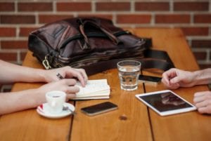 Two people sitting at wooden table with iphones and ipad, coffee and water.